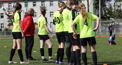 Grupa dziewczyn w strojach piłkarskich stojąca tyłem, w tle bramka do piłki nożnej. - powiększ