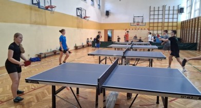 Zdjęcie przedstawia wgląd na całą halę sportową na której rozstawione są stoły do gry w tenisa stołowego.
Dzieci stoją przy stołach i grają. Każdy zawodnik jest bardzo skupiony na swojej grze. - powiększ