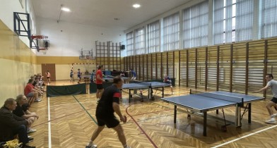 Zdjęcie przedstawia widok na na całą halę sportową, gdzie zawodnicy w każdej kategorii wiekowej zmagają się w swoich rozgrywkach przy stołach do tenisa stołowego i rozgrywają swoje mecze. Na twarzach zawodników widać zaangażowanie i pełne skupienie. - powiększ